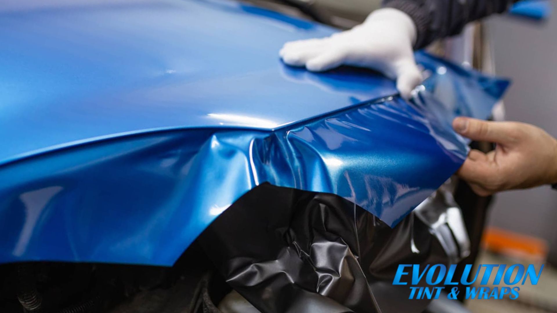 A vibrant blue vinyl wrap being applied to a car’s hood, with hands smoothing the material over the surface. The "Evolution Tint and Wraps" logo is visible in the corner