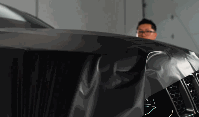 A technician carefully applies a black vinyl wrap onto the hood of a car, showcasing the precision and attention to detail in the wrapping process