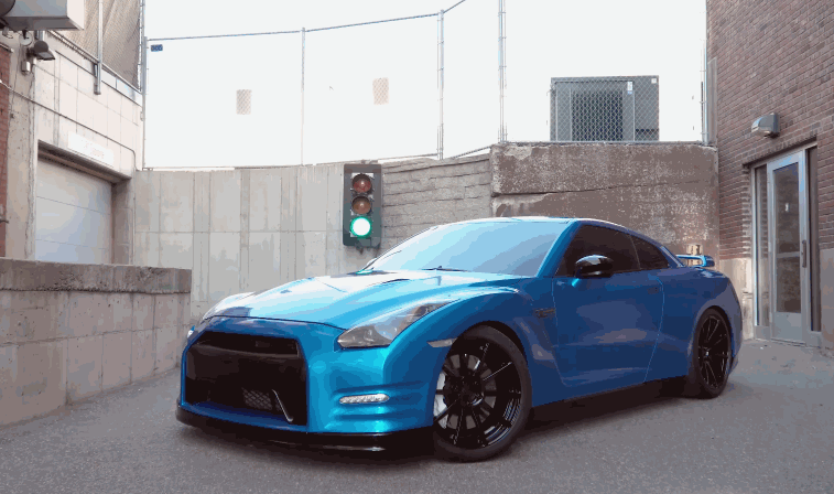 A shiny blue sports car parked outdoors, displaying its glossy vinyl wrap and high-end design with black wheels, under a green traffic light