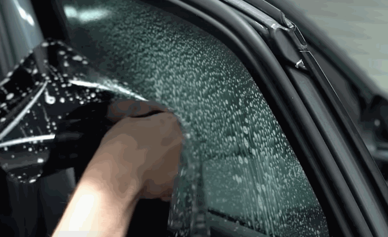 A technician begins applying tinted film to a pre-sprayed car window, aligning it carefully for coverage