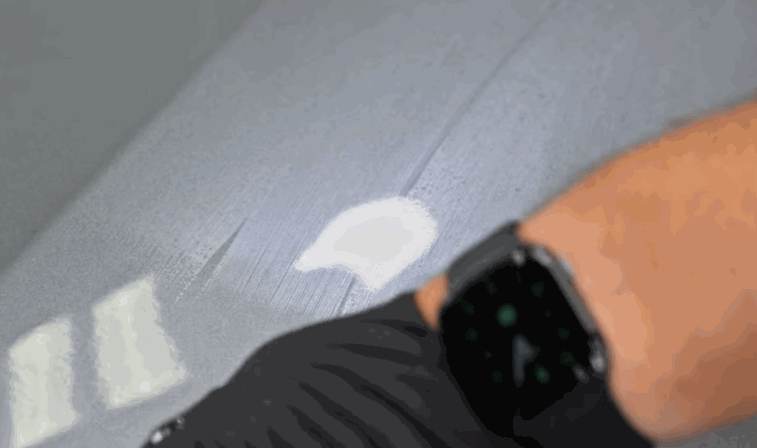 Close-up of a gloved hand spreading white polishing compound across a dull, gray car surface, revealing a smoother finish underneath.