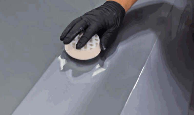 A person in black gloves uses a white, textured foam pad to buff the car’s surface, enhancing the gloss.