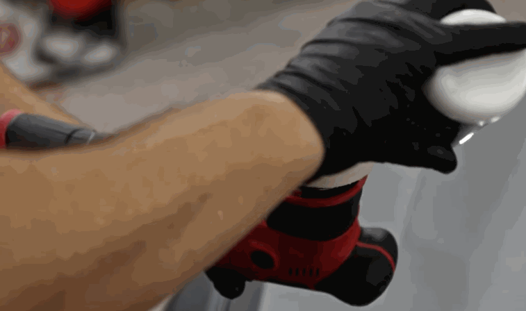 A person wearing black gloves applies a white polishing compound to a red and black dual-action polisher before using it on a car’s surface.