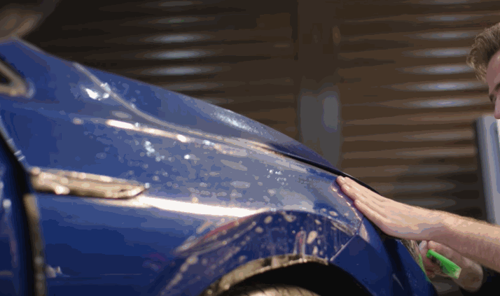 A technician presses down on paint protection film while applying it to the fender of a car, using a green squeegee tool.
