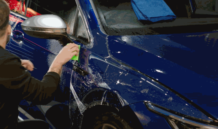 A person uses a green squeegee to smooth out paint protection film on a blue SUV, removing water and air bubbles during vinyl wrap application.