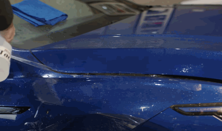 A close-up of a person cutting or aligning protective film on the hood of a blue vehicle, with a blue microfiber cloth nearby.