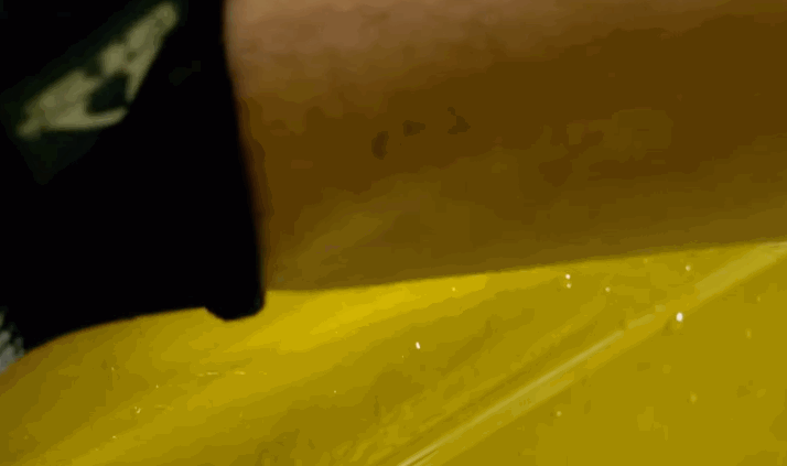 A close-up of a person's hand using a yellow squeegee to apply or smooth out paint protection film on a yellow vehicle surface.