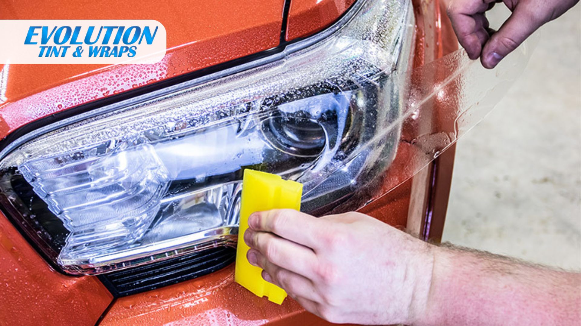 A person applies clear protective film over a headlight of an orange car, using a yellow squeegee to press it into place. The “Evolution Tint & Wraps” logo appears in the top left corner.