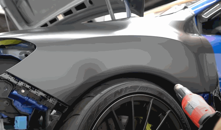 A heat gun is used to stretch matte black vinyl wraps across the rear quarter panel of a sports car, helping the material conform to the curves.