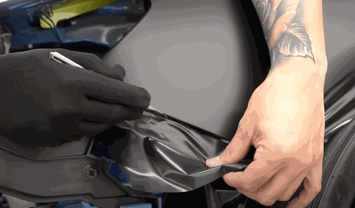 Two hands, one gloved and one tattooed, trim excess matte vinyl wraps along a car panel using a precision blade for a clean edge.