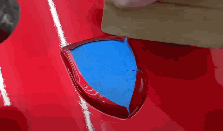 A person precisely applies vinyl wraps into a recessed area of a red car using a flat tool, ensuring the wrap adheres smoothly to the surface.