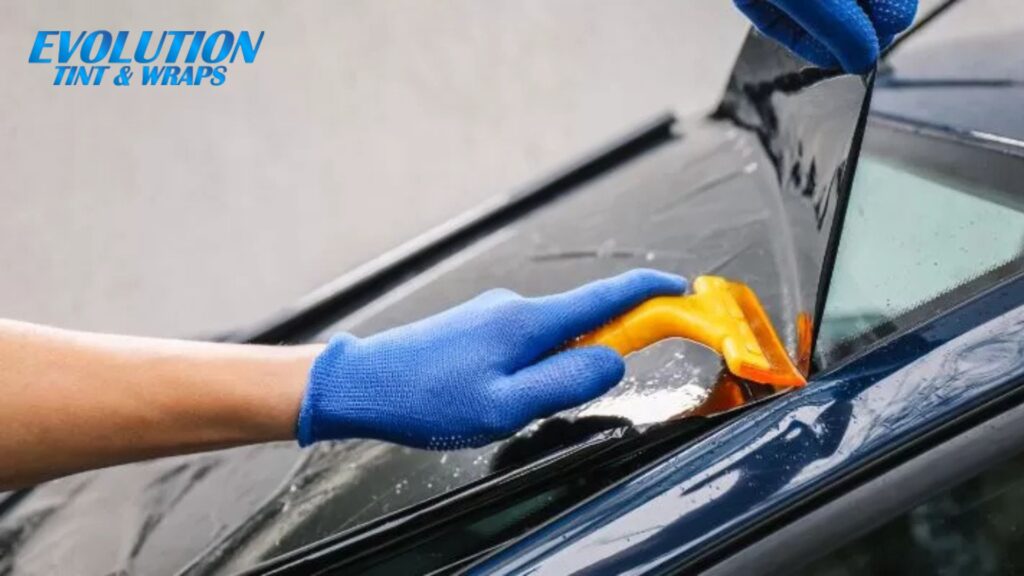 Person wearing blue gloves using a yellow tool to press dark window tint film onto a car window, with the Evolution Tint & Wraps logo in the corner.