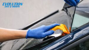 Person wearing blue gloves using a yellow tool to press dark window tint film onto a car window, with the Evolution Tint & Wraps logo in the corner.