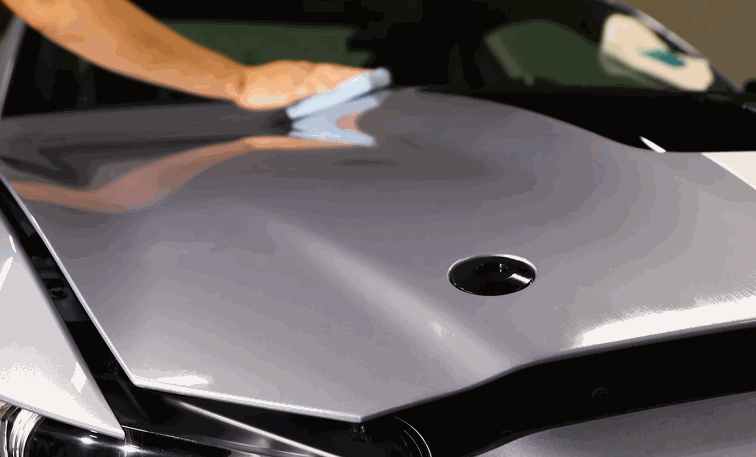 A person uses a microfiber towel to buff the ceramic-coated hood of a silver sports car under bright lighting.