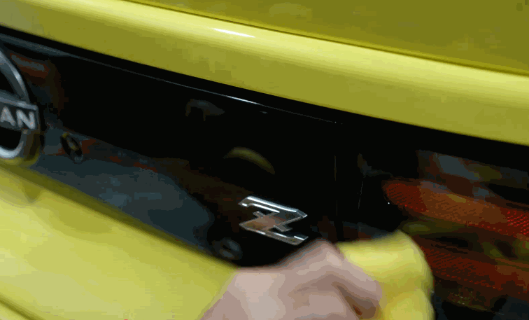 A person buffs the rear panel of a yellow Nissan sports car with a yellow microfiber cloth, highlighting the “Z” emblem and glossy black finish.