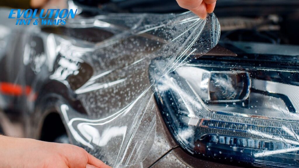 A close-up of a person applying a clear paint protection film over a car headlight and front bumper, with the logo “Evolution Tint & Wraps” in the top left.