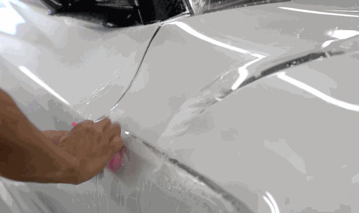 A technician smooths a wet paint protection film onto the rear quarter panel of a white car using a pink squeegee.