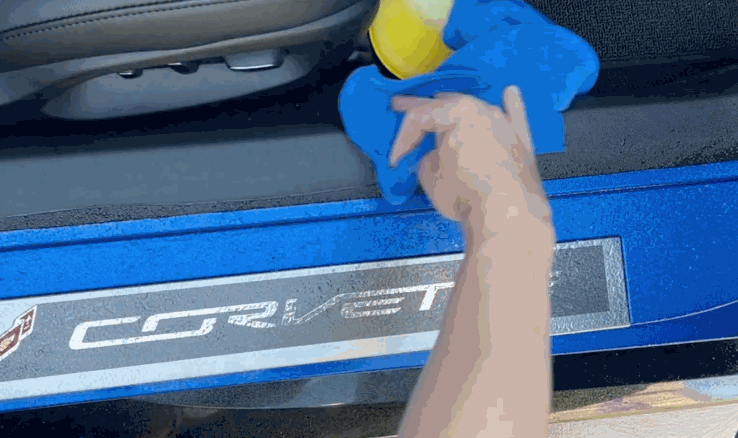 A hand wipes down the inner door sill of a blue Corvette using a blue microfiber towel and a yellow spray bottle.