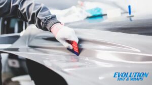 A technician wearing gloves smooths out a vinyl wrap on a car's body using a red and blue squeegee. The wrap is being applied with precision to eliminate air bubbles.
