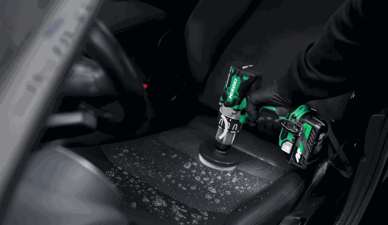 A power scrubber with green accents deep cleans a foam-covered car seat, loosening embedded dirt as part of a car detailing process.