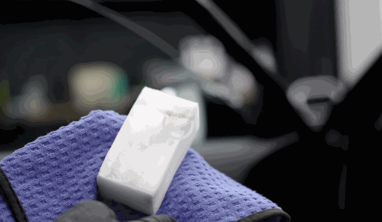 A close-up of a melamine sponge held over a microfiber towel, used to clean scuff marks from interior car panels during detailing.