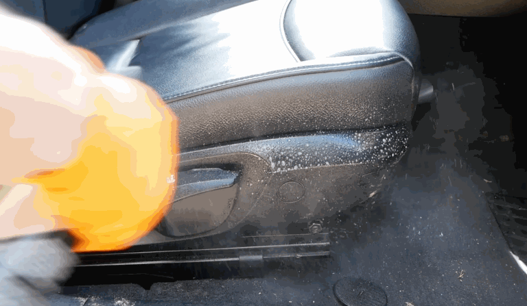 A gloved hand uses an orange microfiber cloth to wipe down the lower side of a car seat, removing dirt and cleaning product foam during interior car detailing.