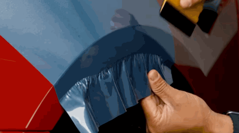 Hands stretch and smooth a blue vinyl wrap over a rounded car edge, carefully removing tension to avoid wrinkles.