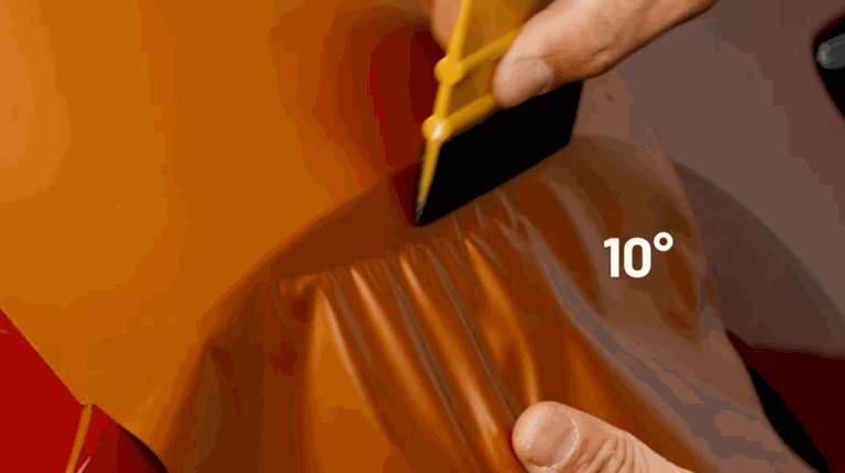 Close-up of a yellow squeegee held at a 10-degree angle, smoothing orange vinyl wrap over a contoured car panel.