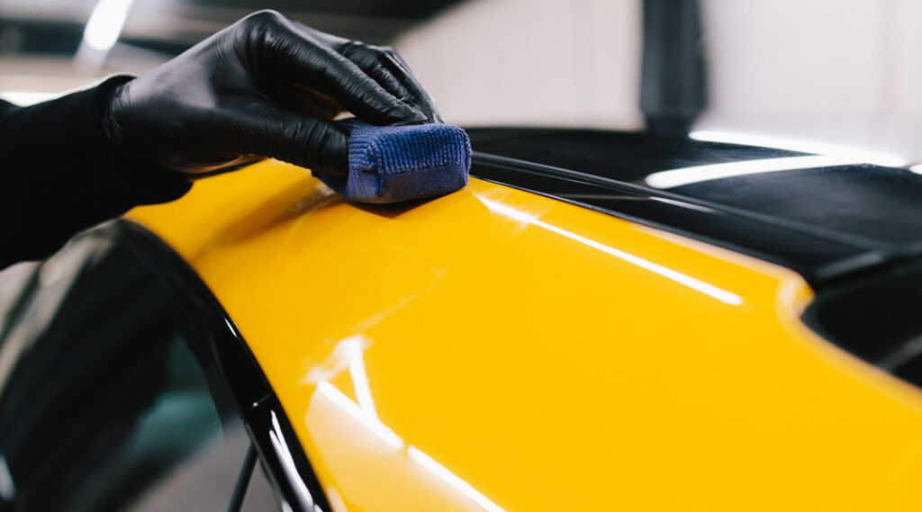 A person wearing black gloves applies ceramic coating to the roof edge of a bright yellow car using a blue microfiber applicator pad.