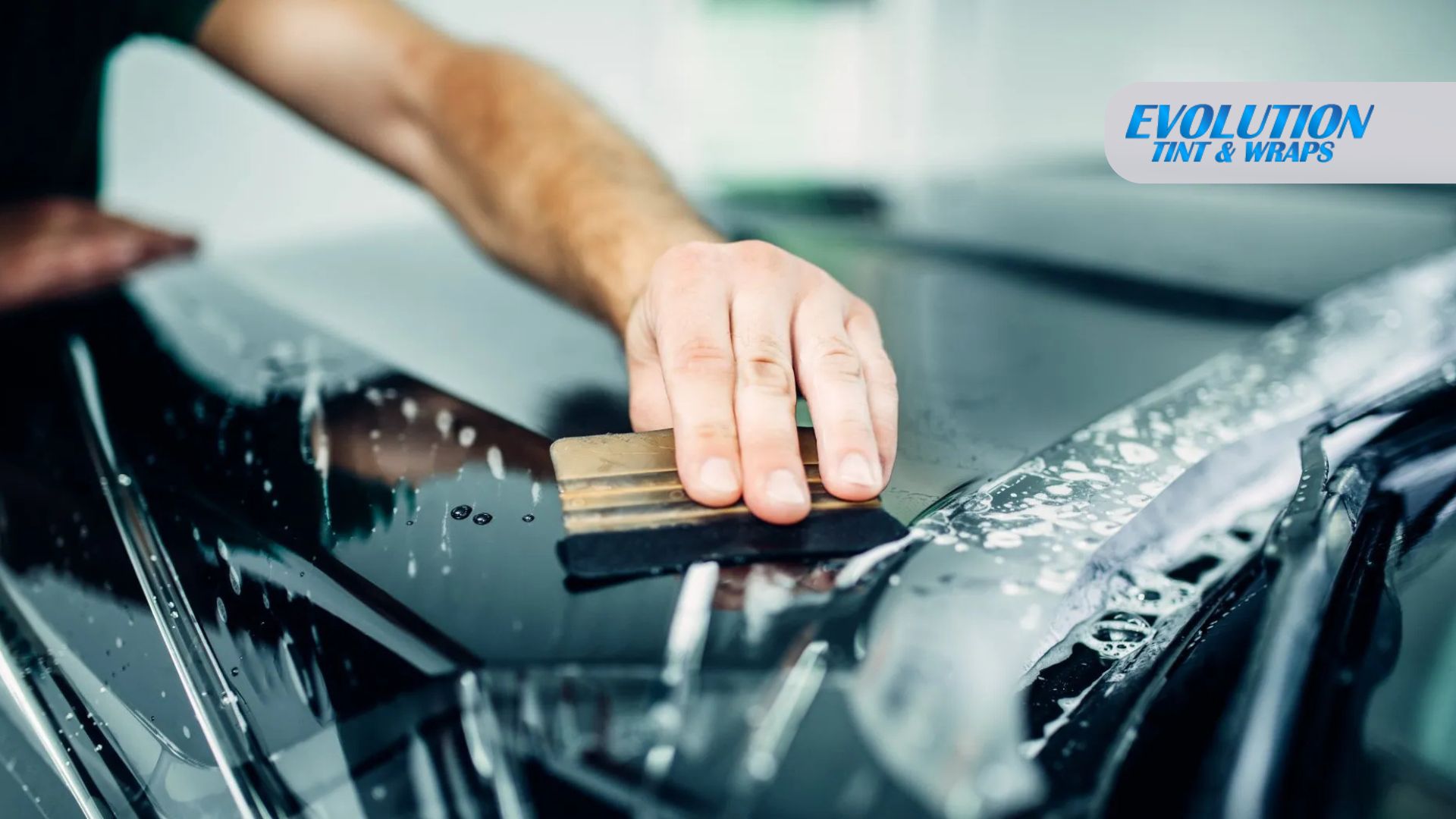 A close-up of a hand using a squeegee to apply tint film on a car windshield, with soap and water being pushed out during the tinting process.