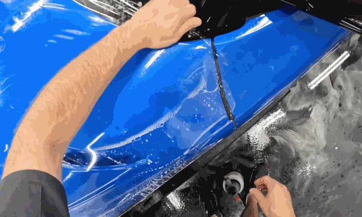 Hands press and adjust a sheet of clear protective film onto the side panel of a glossy blue vehicle. The surrounding area is wet from installation solution.