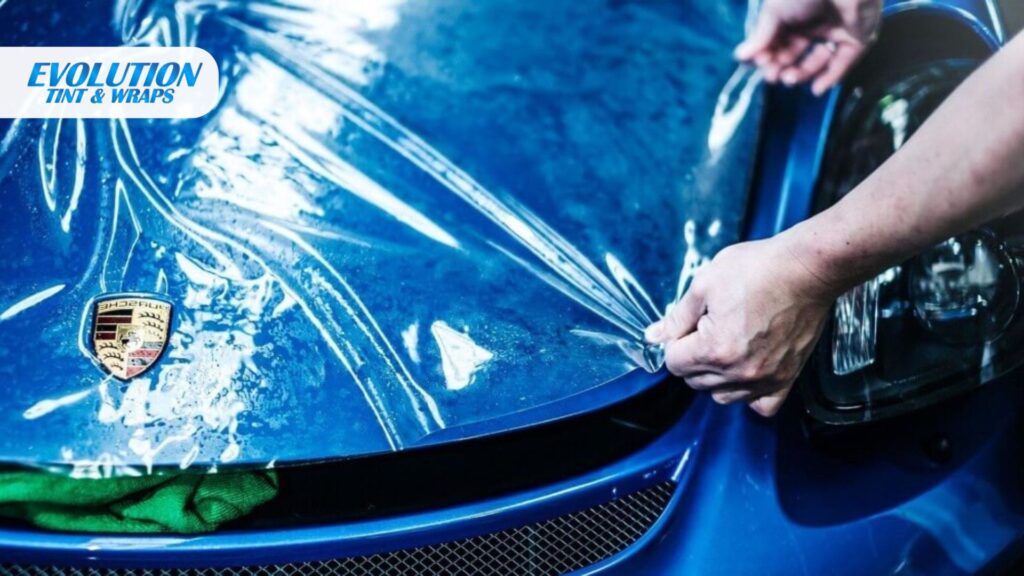 A close-up of a clear film being pulled over the hood of a blue Porsche with a visible logo, with the Evolution Tint & Wraps logo in the top-left corner.