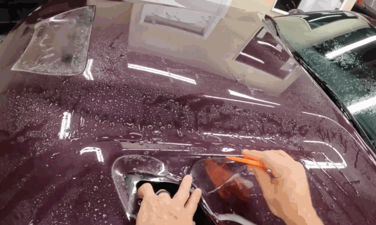 A worker uses a plastic squeegee tool to install clear paint protection film around the hood vent of a deep maroon car. Water droplets cover the surface.