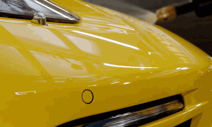 A yellow car’s front bumper and hood are shown under bright lights with a glossy, just-installed paint protection film being smoothed out.