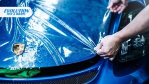 A close-up of a clear film being pulled over the hood of a blue Porsche with a visible logo, with the Evolution Tint & Wraps logo in the top-left corner.