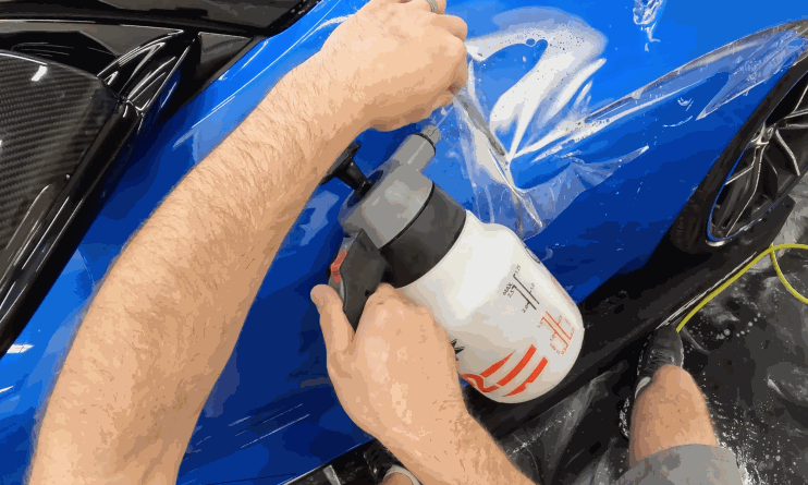 A person sprays solution onto the fender of a blue car while applying a clear paint protection film, smoothing it out with their hands.