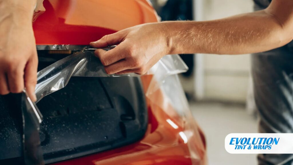 Person carefully stretches and aligns clear protective film over the rear end of an orange car.