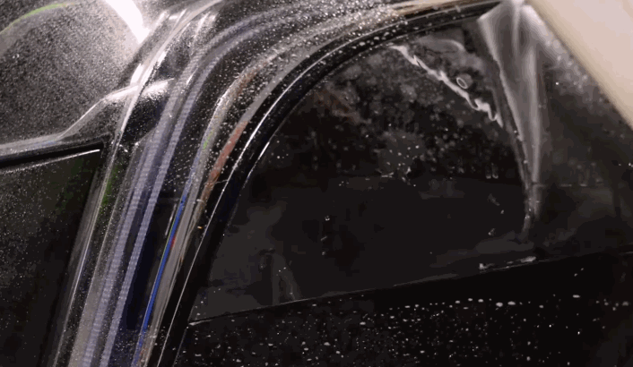 Transparent film being pulled tightly over a glossy black car surface with water droplets.