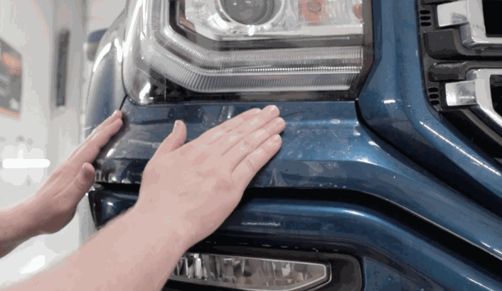 Hands smoothing paint protection film onto the blue front bumper and headlight of a truck.