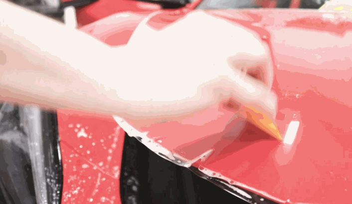 Installer uses a yellow squeegee to smooth clear film over the curved red hood of a vehicle.