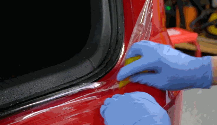 A person wearing blue gloves uses a yellow squeegee to apply paint protection film (PPF) around the rear window of a red car.