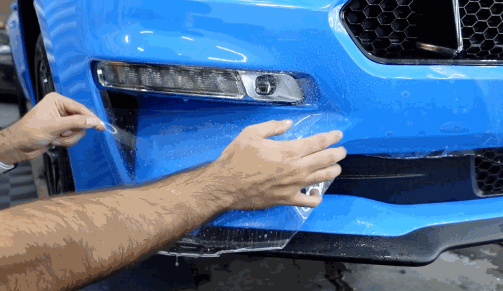 Hands carefully stretch and press clear paint protection film onto the front bumper of a blue car, ensuring a precise fit around curves and edges.