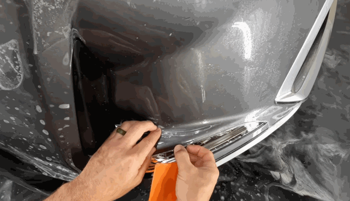 A person smooths paint protection film over the headlight area of a grey car using an orange squeegee while the surface is still wet.
