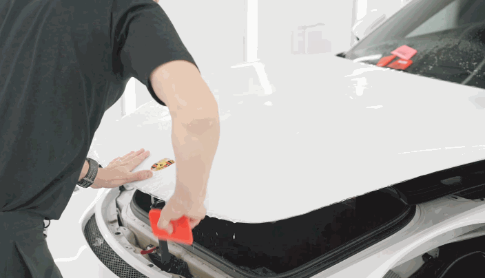 A person applies paint protection film on the hood of a white Porsche, using a red squeegee to remove bubbles and ensure a smooth finish.