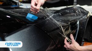 A close-up of hands stretching and positioning clear paint protection film over the hood and fender of a black car
