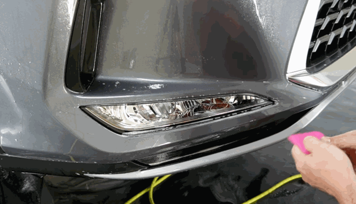 A close-up of hands using a pink squeegee to apply paint protection film to the bumper of a wet grey vehicle.