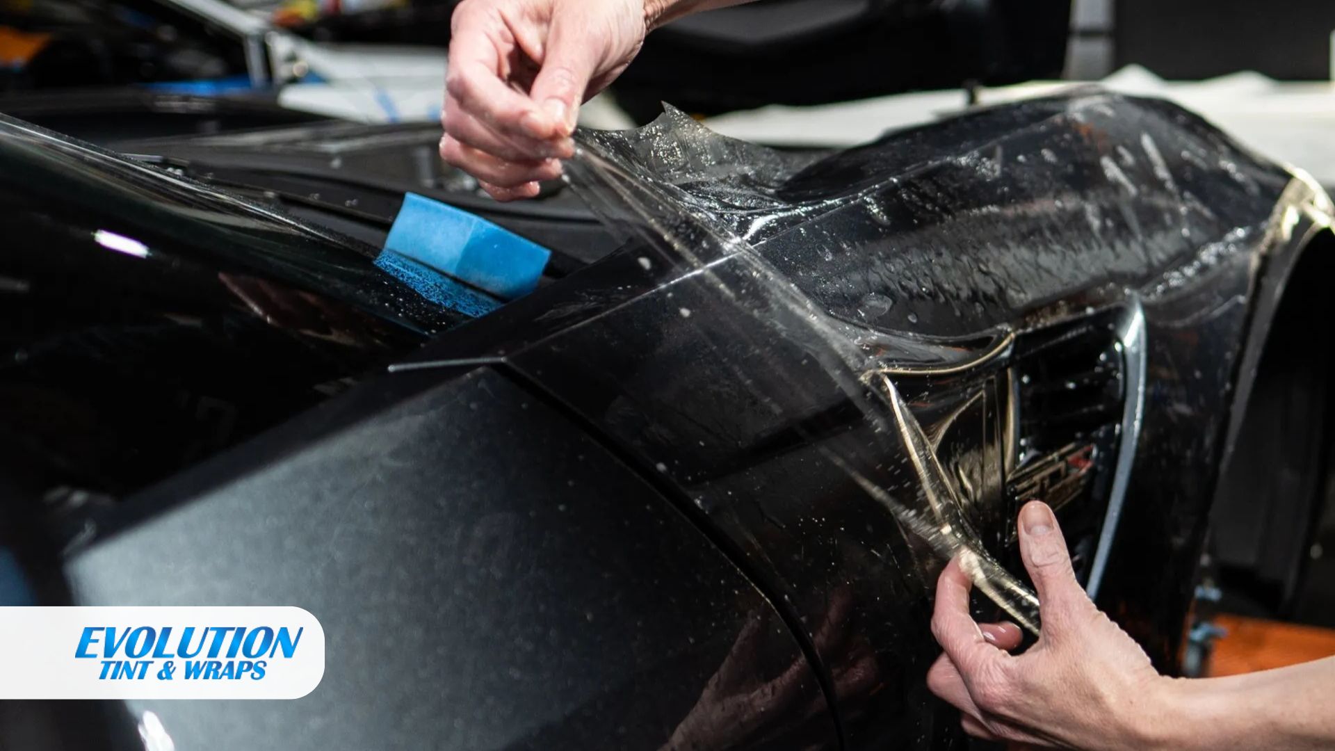A close-up of hands stretching and positioning clear paint protection film over the hood and fender of a black car