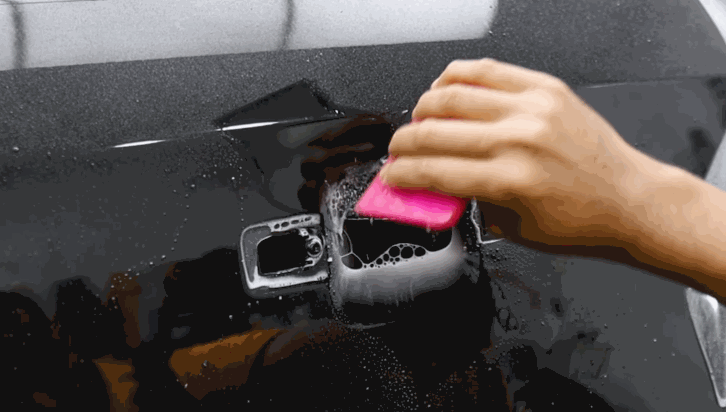 A person smooths out paint protection film on a glossy black car panel using a pink squeegee, focusing around a recessed area.