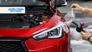 A technician applies paint protection film to the headlight and front fender of a red car, using a spray bottle to smooth the film.