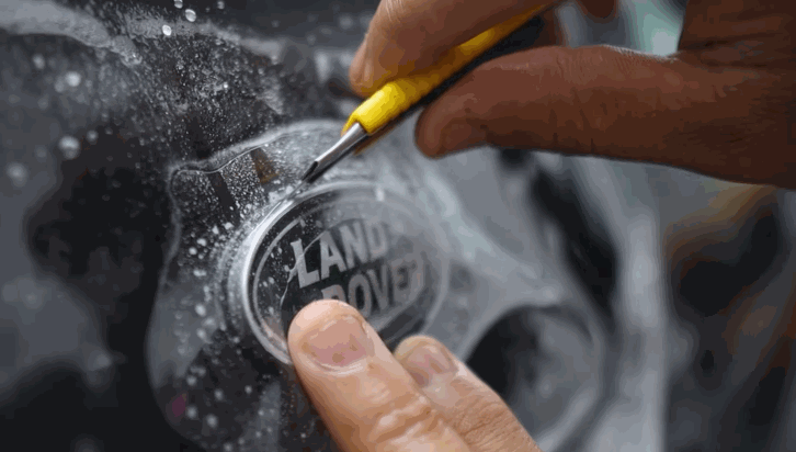 Close-up of a technician using a precision knife to trim paint protection film around a Land Rover badge on a wet car surface.